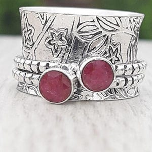 May include: A silver ring with a floral design and two round, red gemstones. The ring has a textured band and a decorative top with etched leaves and star-shaped cutouts. The gemstones are a deep red color.