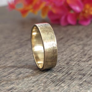 May include: A gold-coloured metal ring with a flat band and a textured, brushed finish on the exterior surface. The interior of the band is smooth and polished, reflecting light, while the outer surface shows fine, vertical scratch-like markings.