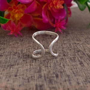 May include: A silver ring with a unique open design. The ring features a curved shape that wraps around the finger, with a small gap in the center. The ring is set against a wooden surface with pink and orange flowers in the background.