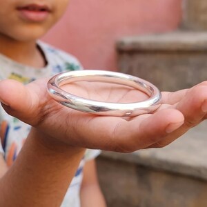 May include: A smooth, polished silver-toned bangle bracelet with a rounded, circular profile. The metal surface is highly reflective, showing light highlights along the curved edges of the solid, continuous band design.