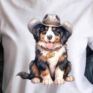 Op de afbeelding: Wit T-shirt met een aquarel illustratie van een Berner Sennenhond met een cowboyhoed. De hond heeft zwart, bruin en wit haar, een bruine halsband en een gouden hanger. De hond lacht met zijn tong uit.