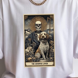 May include: White t-shirt featuring a tarot card-style graphic. The design includes a skeleton with a halo, a Cavapoo dog, and the text "THE CAVAPOO LOVER" in a gold frame. The artwork has a vintage aesthetic.