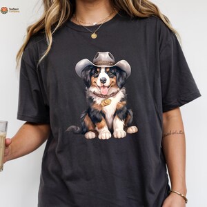 Op de afbeelding: Donkergrijs T-shirt met een aquarel illustratie van een Berner Sennenhond met een cowboyhoed. De hond heeft een bruin-zwarte vacht, een gouden hanger en lacht met zijn tong uit. Het shirt is een comfortabele, casual stijl.