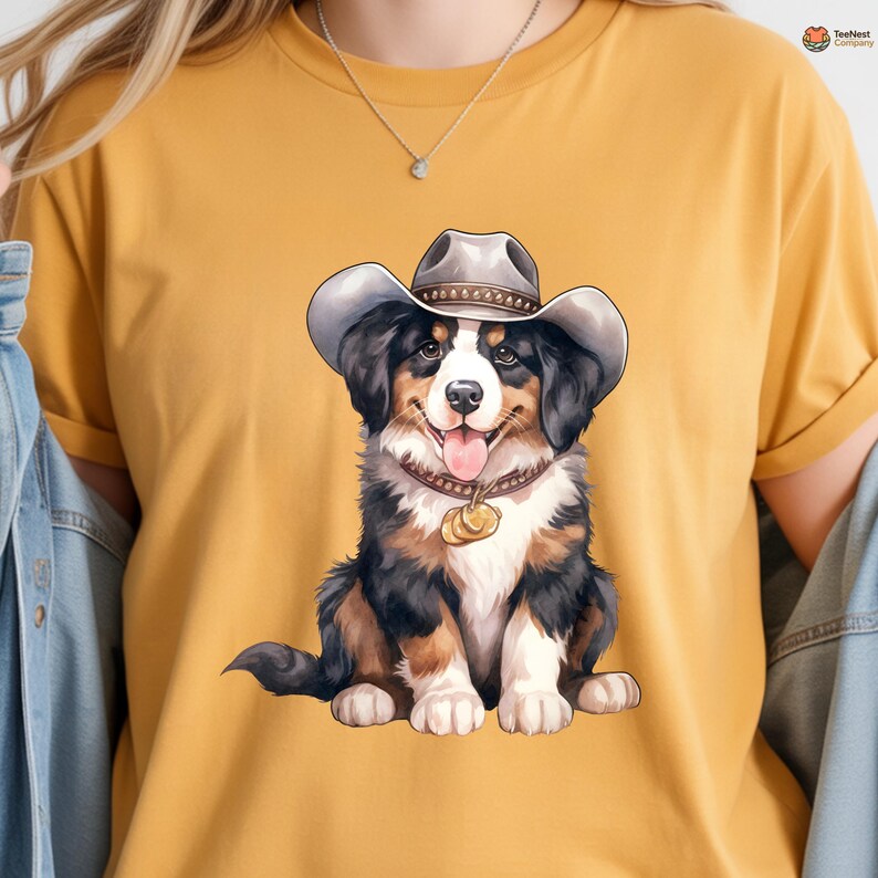 Op de afbeelding: Mosterdgeel T-shirt met een cartoon illustratie van een Berner Sennenhond met een cowboyhoed. De hond heeft zwart, bruin en wit haar, een bruine halsband met een gouden hanger en zijn tong steekt uit.