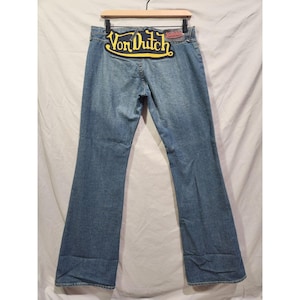 May include: Light blue denim jeans with a flared leg. The jeans feature a large yellow and black Von Dutch patch on the back waistband. A red Von Dutch label is also visible. The jeans are hanging on a wooden hanger.