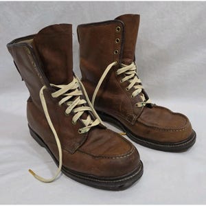 Op de afbeelding: Een paar bruine leren veterschoenen met een mocassin-neus, crèmekleurige veters en messingkleurige oogjes. De laarzen hebben een hoog model met zichtbare stiksels op de neus en een donkerbruine zool met een hak.