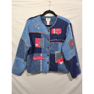 May include: A patchwork denim jacket in shades of blue, featuring heart and floral appliqués. The jacket has a button-up front and a V-neck collar. Decorative stitching and patches add to the unique, handcrafted look. The jacket is hanging on a black hanger.