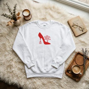 May include: White sweatshirt featuring a red high-heeled shoe graphic with the text "THE DEVIL WEARS PRADA 2". The design includes a trident. The sweatshirt is displayed on a fluffy, white surface.