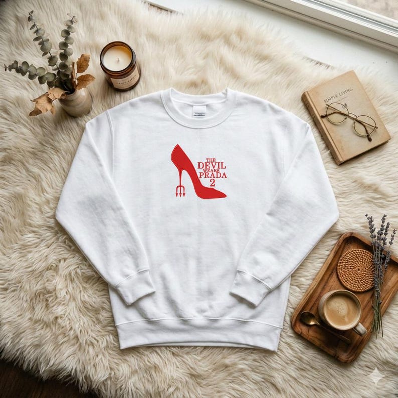 May include: White sweatshirt featuring a red high-heeled shoe graphic with the text "THE DEVIL WEARS PRADA 2". The design includes a trident. The sweatshirt is displayed on a fluffy, white surface.