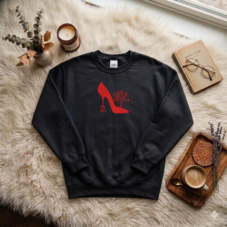 May include: Black sweatshirt with a red high-heeled shoe graphic and the text "The Devil Wears Prada 2". The shoe has a trident heel. The sweatshirt is displayed on a fluffy white surface.