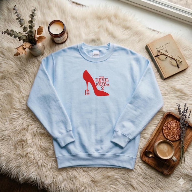 May include: A light blue sweatshirt featuring a red high-heeled shoe design with the text "THE DEVIL WEARS PRADA 2". The sweatshirt is displayed on a fluffy white rug, with a candle, book, and coffee cup in the background.