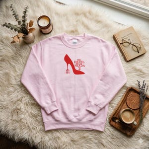 May include: A light pink sweatshirt with a red high heel and the text "THE DEVIL WEARS PRADA 2". The design includes a red trident. The sweatshirt is on a fluffy white surface.