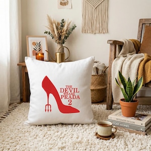 May include: White square throw pillow featuring a red high-heeled shoe design with a trident heel and the text "THE DEVIL WEARS PRADA 2". The pillow is placed on a white rug, suggesting a home decor theme.