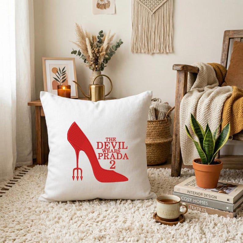 May include: White square throw pillow featuring a red high-heeled shoe design with a trident heel and the text "THE DEVIL WEARS PRADA 2". The pillow is placed on a white rug, suggesting a home decor theme.