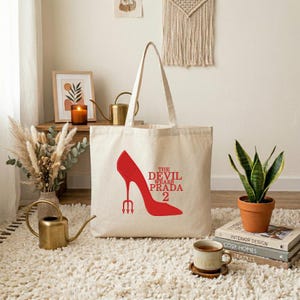 May include: A cream-colored tote bag featuring a red high-heeled shoe design with a trident and the text "THE DEVIL WEARS PRADA 2". The bag is made of a sturdy fabric and has two handles for carrying.