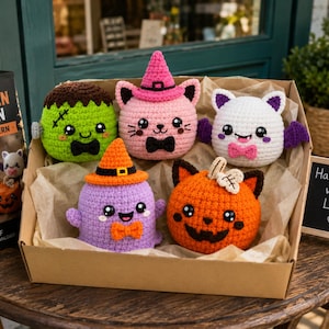 Puede incluir: Un conjunto de cinco peluches de ganchillo con temática de Halloween en una caja de cartón, que incluye un monstruo verde, un gato rosa con sombrero de bruja, un murciélago blanco, un fantasma morado y una calabaza naranja.