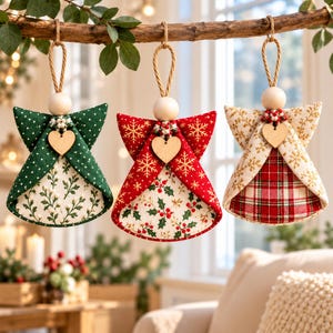 Peut inclure: Trois décorations d'anges en tissu suspendues à une branche. L'ange de gauche est vert à pois blancs avec un motif floral. L'ange central est rouge avec des flocons dorés et du houx. L'ange de droite présente un motif flocon crème et un tartan rouge.