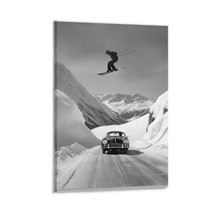 May include: Black and white print of a vintage car driving on a snowy road, with a skier mid-air in the background. The scene is set against a backdrop of snow-covered mountains and a cloudy sky, evoking a sense of adventure.