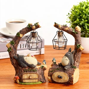 May include: Two decorative lamps featuring a tree trunk design with a cartoon character on each. Each lamp has a light bulb in a black wire cage hanging from a branch. The lamps are brown, green, and beige.
