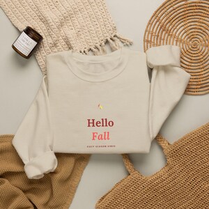 Könnte beinhalten: Ein cremefarbenes Sweatshirt mit Rundhalsausschnitt und dem Schriftzug "Hello Fall" in roten und rosa Buchstaben, darunter der kleinere Text "COZY SEASON VIBES". Über dem Wort "Hello" befindet sich eine kleine, bunte Blattgrafik.