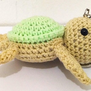 May include: A handmade crochet turtle keychain. The turtle has a light green shell and a tan body, head, and limbs. It features a black button eye and a metal keyring. The turtle is a cute, whimsical accessory.