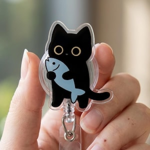 Puede incluir: Un portatarjetas retráctil transparente con un gato negro sosteniendo un pez azul. El gato tiene ojos grandes y redondos y una expresión sonriente. El portatarjetas tiene un clip de plástico transparente y un cierre de metal plateado.