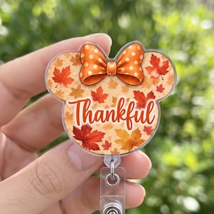 Puede incluir: Un portatarjetas con silueta de orejas de ratón y un patrón de hojas de arce otoñales en naranja y rojo. Un lazo naranja con lunares blancos se sitúa en la parte superior. La palabra Thankful aparece en cursiva naranja en el centro.