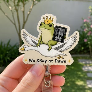 May include: A badge reel featuring a die-cut acrylic topper of a green frog wearing a gold crown and holding a black X-ray film while riding on the back of a white goose. The text We XRay at Dawn is printed in black below the illustration.