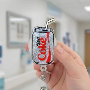 May include: A Diet Coke badge reel with a silver glitter background and red lettering. The badge reel is in the shape of a Diet Coke can with a white straw. The reel has a clear plastic strap and a metal clip.