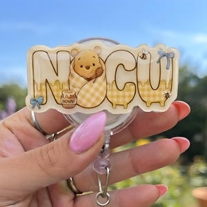 May include: A circular badge reel featuring a custom acrylic topper with the letters N, O, C, U in a yellow and white checkered pattern. The design includes a cartoon bear wrapped in a blanket, a honey pot, small bees, and blue bows.