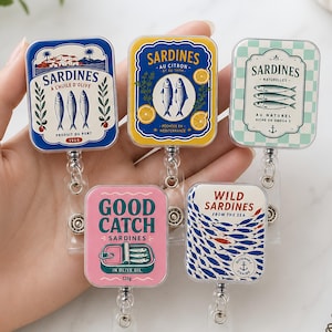 Op de afbeelding: Vijf rechthoekige badgehouders met sardine-ontwerpen. De bovenste rij bevat een blauw blik met drie vissen, een geel blik met twee vissen en citroenschijfjes, en een groen geblokt blik. De onderste rij toont een roze blik met de tekst Good Catch en een wit blik met een school blauwe vissen.