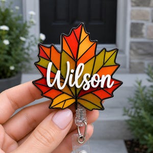 May include: A maple leaf-shaped badge reel featuring a stained-glass style design with vibrant red, orange, and yellow segments outlined in black. The name Wilson is written across the center in white, raised, cursive script lettering.