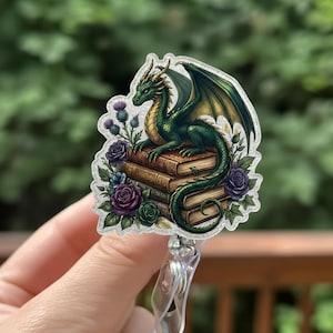 May include: A badge reel featuring a green dragon with gold wings perched atop a stack of books. The dragon is surrounded by purple and blue roses and thistles. The reel has a clear plastic clip.