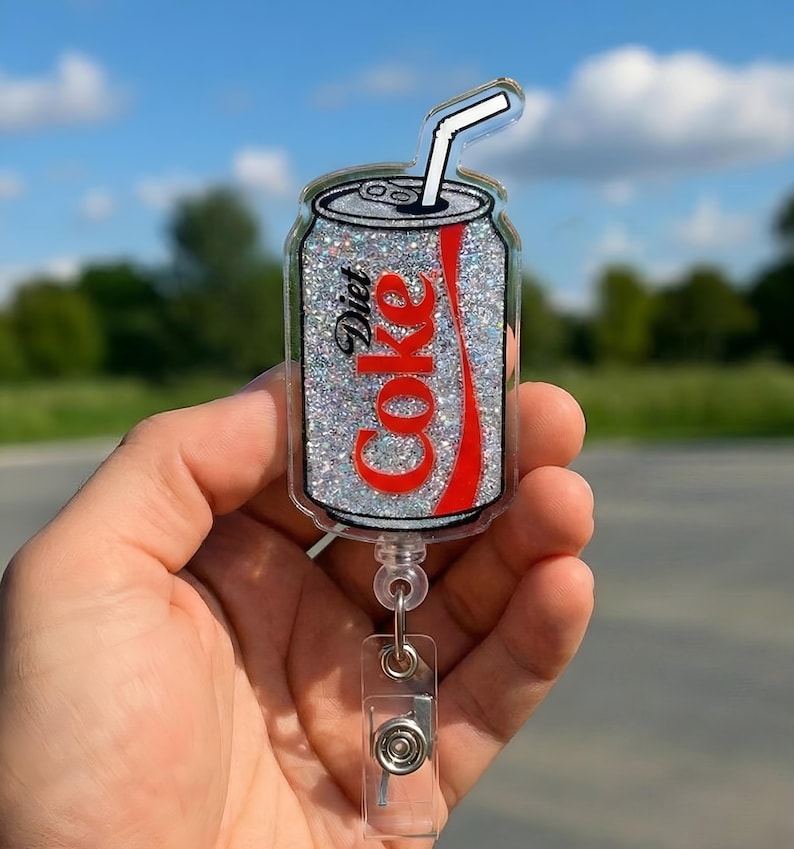 May include: A Diet Coke can-shaped badge reel with a glittery silver background and the Diet Coke logo in red. The badge reel has a clear plastic retractable cord and a clear plastic clip.