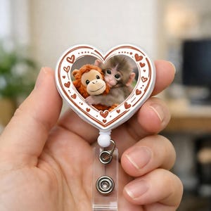 May include: A heart-shaped retractable badge holder featuring a photo of two monkeys hugging. The white border is decorated with brown hearts. The badge reel has a clear plastic strap and a white button.