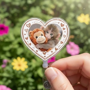 May include: A heart-shaped retractable badge holder featuring a photo of two monkeys hugging. The badge holder has a white background with a brown border of hearts. The background is a blurred image of green foliage and colorful flowers.