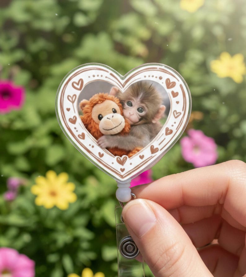 May include: A heart-shaped retractable badge holder featuring a photo of two monkeys hugging. The badge holder has a white background with a brown border of hearts. The background is a blurred image of green foliage and colorful flowers.