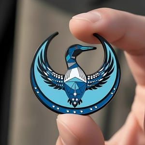 May include: A round, blue and black enamel pin featuring a stylized bird design. The bird has geometric patterns in shades of blue, white, and black, with a red eye. The pin is held between two fingers.