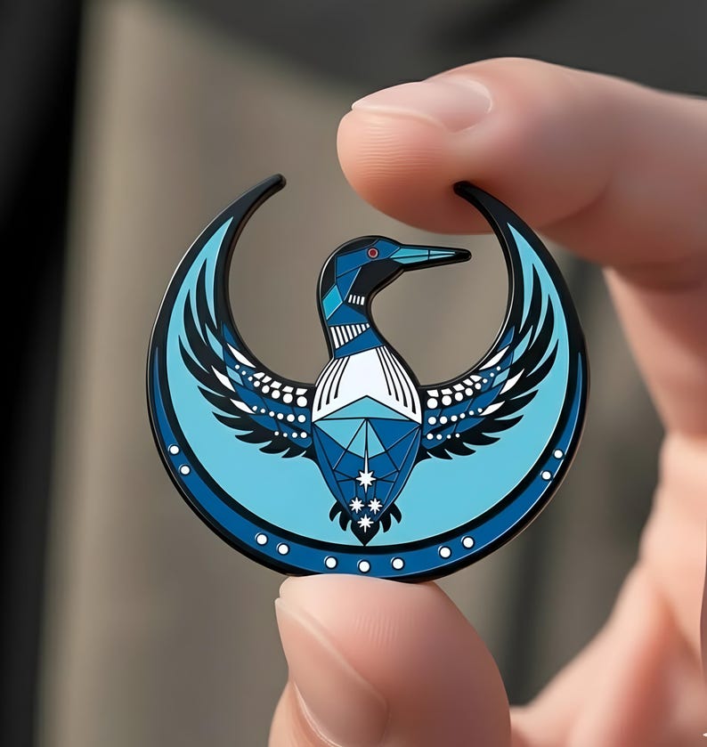 May include: A round, blue and black enamel pin featuring a stylized bird design. The bird has geometric patterns in shades of blue, white, and black, with a red eye. The pin is held between two fingers.