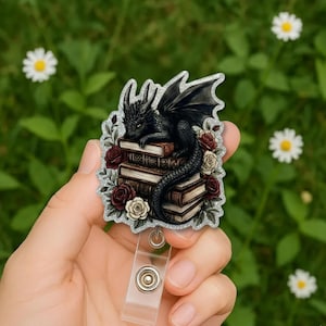 May include: A badge reel featuring a black dragon resting on a stack of books, surrounded by red and cream roses. The dragon has detailed scales and wings. The reel has a clear plastic strap and a silver-colored metal clip.