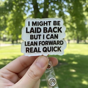 Puede incluir: Un portatarjetas blanco brillante con el texto "I MIGHT BE LAID BACK BUT I CAN LEAN FORWARD REAL QUICK" en negro. El portatarjetas tiene una correa de plástico transparente y un clip de metal plateado.