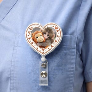 May include: A heart-shaped retractable badge holder featuring two plush monkey toys hugging each other. The badge reel has a white border with small brown hearts. The badge is attached to a clear plastic clip and a metal reel.