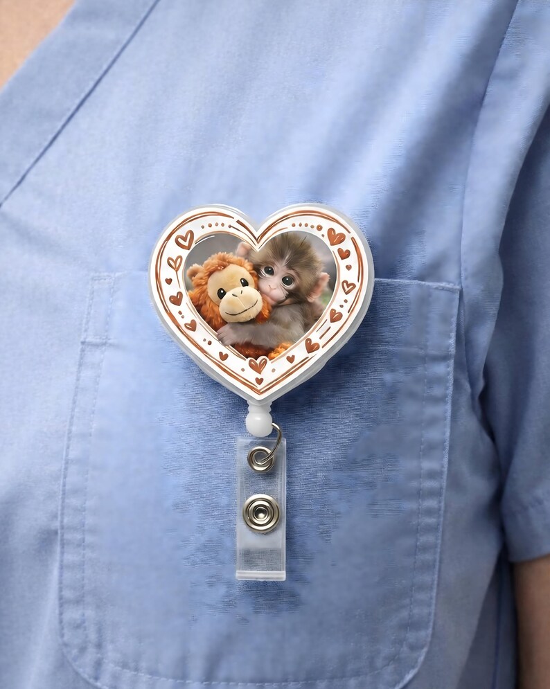 May include: A heart-shaped retractable badge holder featuring two plush monkey toys hugging each other. The badge reel has a white border with small brown hearts. The badge is attached to a clear plastic clip and a metal reel.