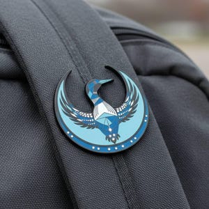 May include: A round, blue and black enamel pin featuring a stylized bird design. The pin is attached to a gray backpack. The bird has geometric shapes and a crescent moon shape around it. The pin has a white border with small white dots.