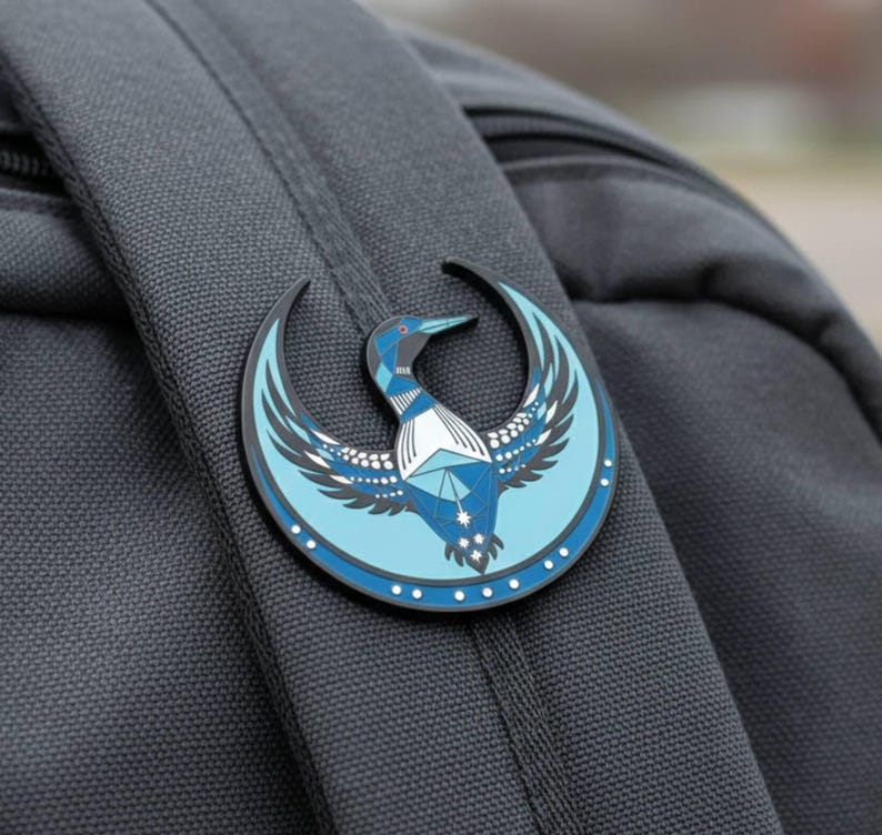 May include: A round, blue and black enamel pin featuring a stylized bird design. The pin is attached to a gray backpack. The bird has geometric shapes and a crescent moon shape around it. The pin has a white border with small white dots.