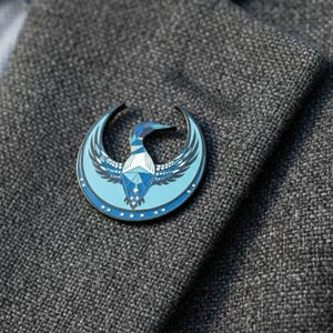 May include: A round, blue and silver enamel pin featuring a stylized loon design. The pin is set against a gray textured fabric, showcasing the bird's wings and geometric body. The loon has a red beak and is surrounded by a crescent shape.