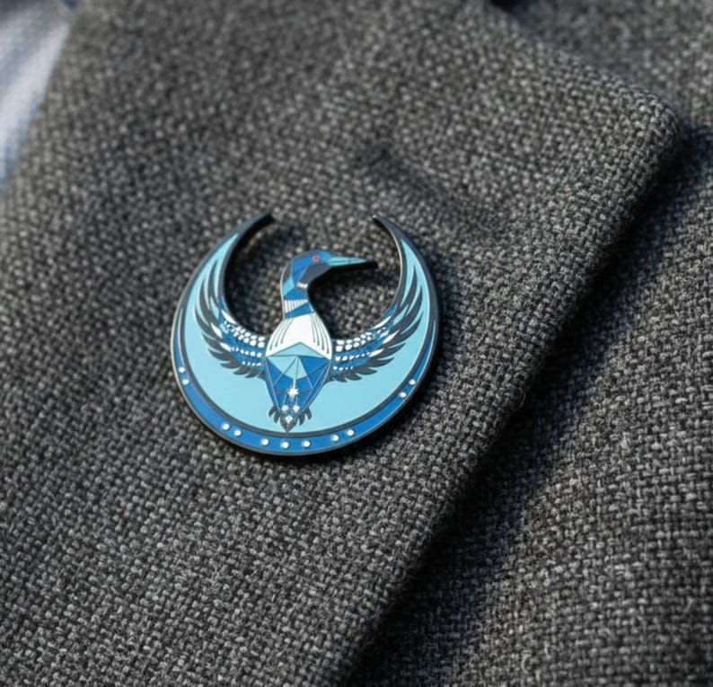 May include: A round, blue and silver enamel pin featuring a stylized loon design. The pin is set against a gray textured fabric, showcasing the bird's wings and geometric body. The loon has a red beak and is surrounded by a crescent shape.
