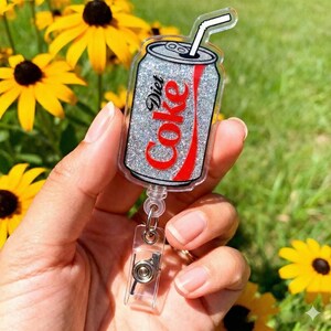 May include: A Diet Coke badge reel shaped like a soda can, featuring a silver glitter effect and a red Coke logo. The reel has a clear plastic retractable cord and a clip for attaching to clothing.