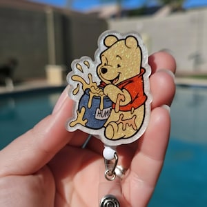 May include: A glittery badge reel featuring a cartoon bear in a red shirt, holding a blue pot of honey with the word "HUM" on it. The badge reel has a clear plastic base and a retractable cord.