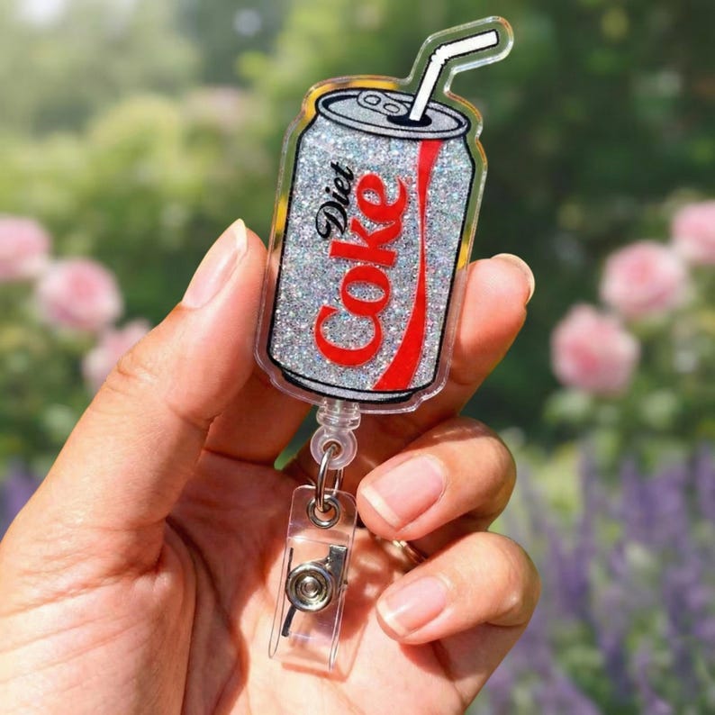 May include: A Diet Coke can-shaped badge reel with a glittery silver background and red Coke logo. The badge reel has a clear plastic clip and a white straw. The text "Diet Coke" is visible on the can.
